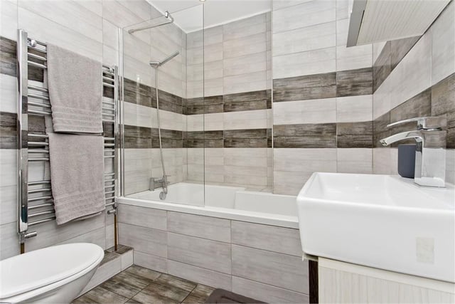 The stunning bathroom features striking contemporary tiles and consists of storage-set washbasin, back-to-the-wall toilet, chrome towel radiator, vanity mirror with storage, and a bathtub with overhead shower and glazed screen.
